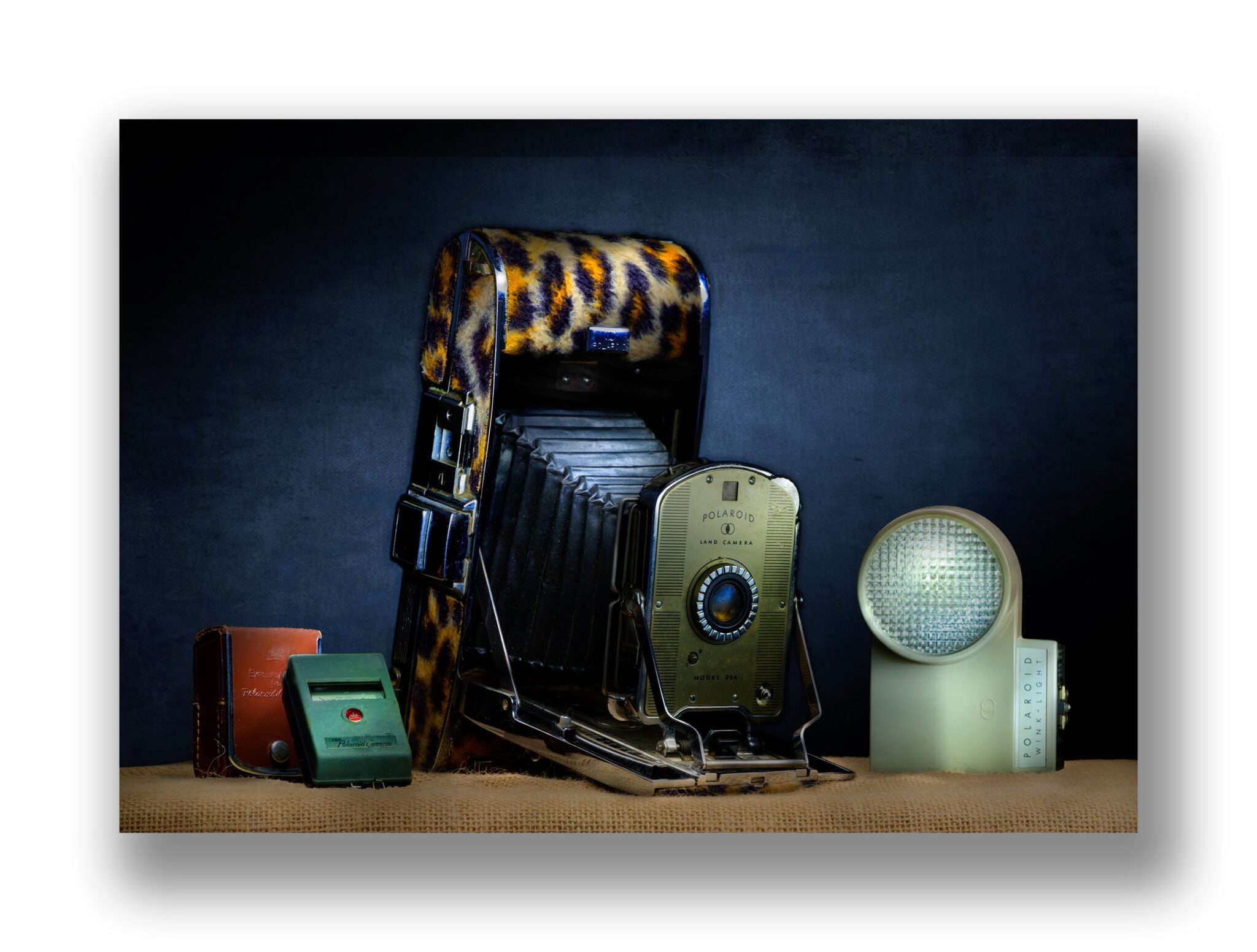 Still life of a vintage Polaroid Land Camera Model 95A with a leopard print case, a Polaroid Wink-Light, and a brown leather case on a burlap surface against a dark blue background.