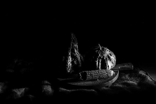 Black-and-white still life light painting featuring gourds and an ear of corn arranged on a wooden surface against a dark background