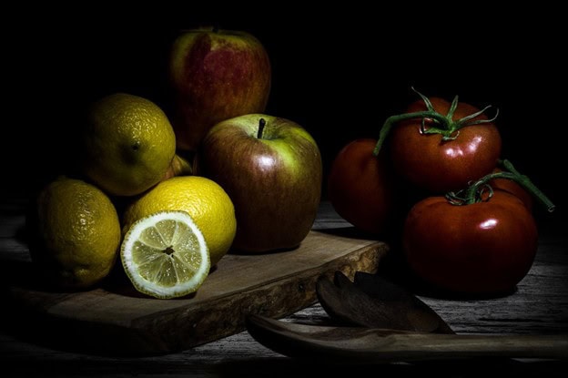 Still life light painting featuring apples, lemons, tomatoes, and a sliced lemon arranged on a wooden surface against a dark background