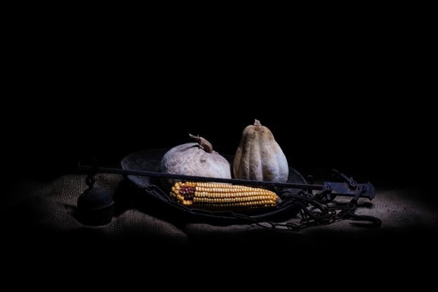 Still life light painting featuring two textured gourds and an ear of corn arranged on a wooden cutting board against a dark background