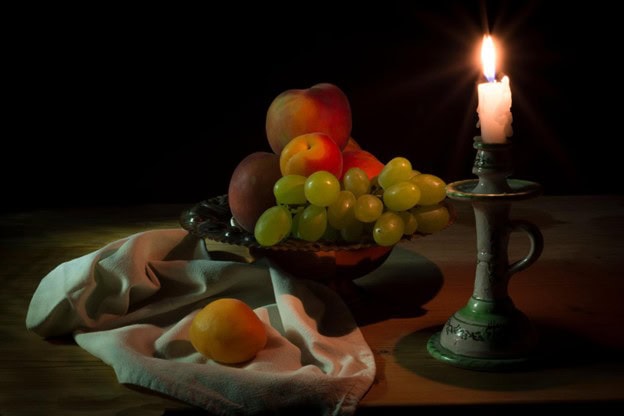Still life photograph featuring grapes, peaches, lemons, and a candle arranged on a table against a dark background