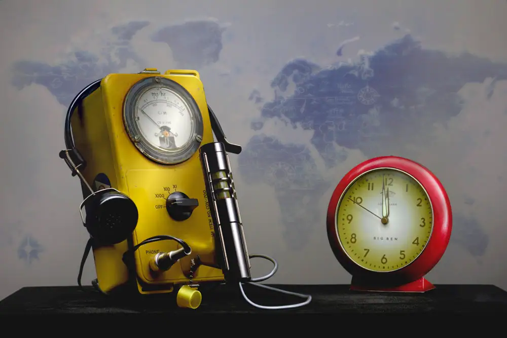 Still-life light painting titled 71 Seconds to Midnight featuring a vintage yellow Geiger counter with headphones beside a red Big Ben alarm clock set near midnight, illuminated against a faded world map background. The composition symbolizes nuclear anxiety, global instability, and the fragility of time.