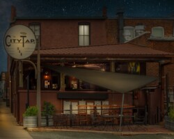 The City Tap Bar is closed for the night, the tables and chairs are empty, and the stars are out. “The City Tap Bar nightscape in Pittsboro, North Carolina illuminated by warm lamplight.”